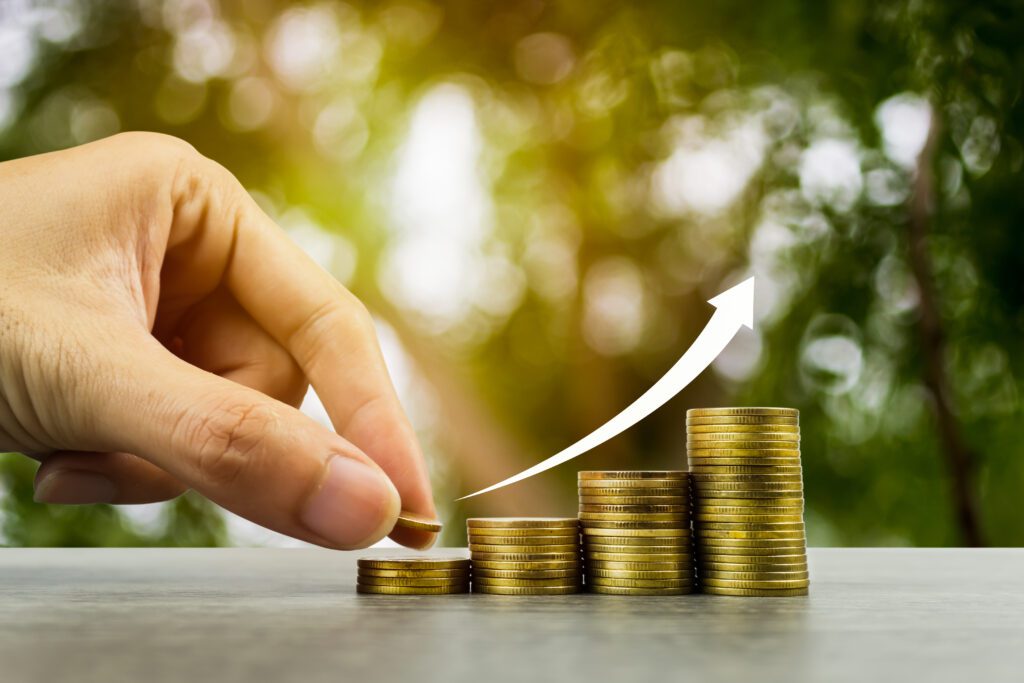 Making money and money investment concept. A man hand holding coin over stack of coins with rise up white arrow on wooden table. Depicts long-term investment And wealth and financial stability.